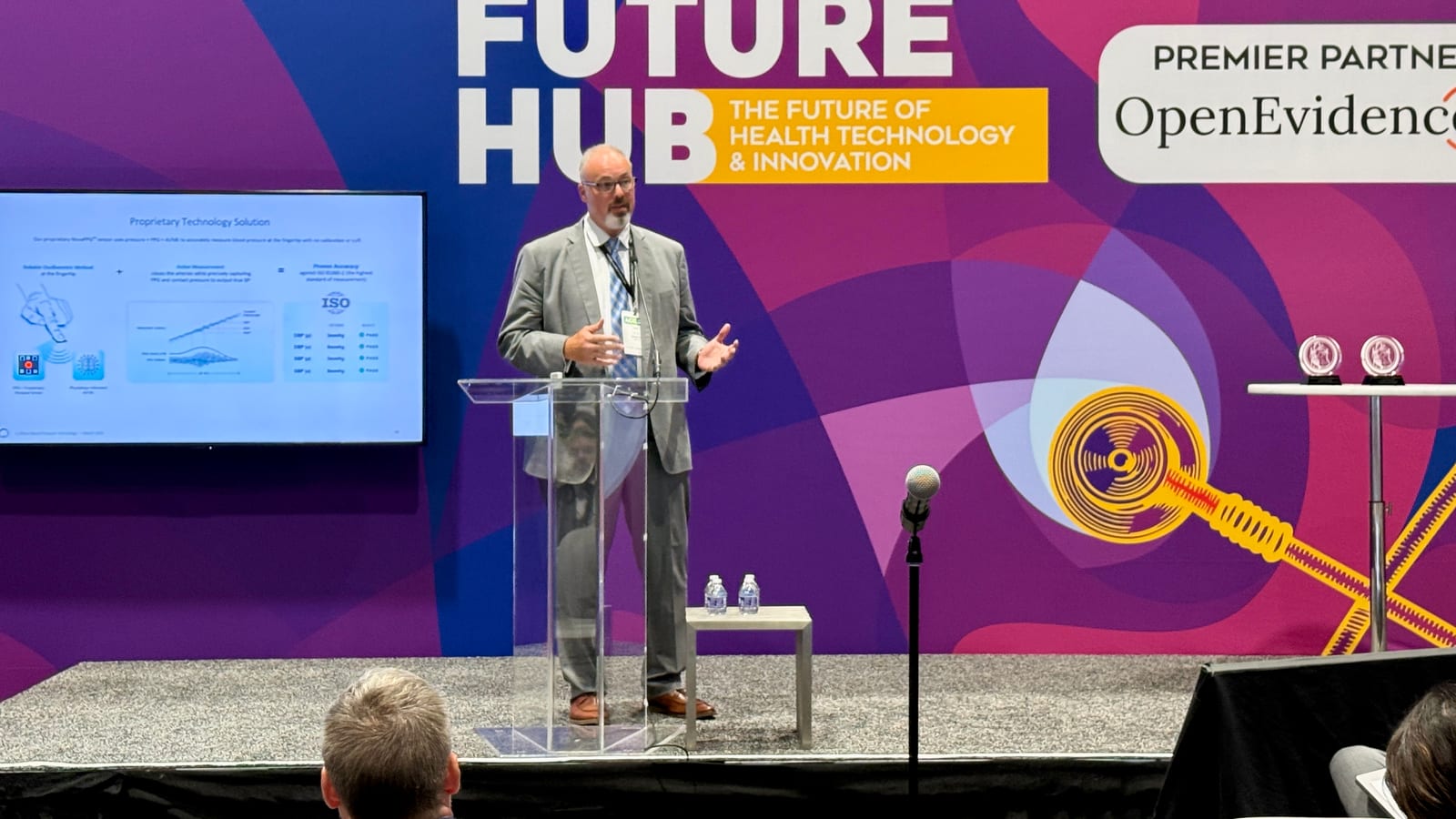 Omer Inan speaks from a clear lectern on the purple Future Hub stage, beside a screen showing a Biozen technology slide, with audience members in the foreground.